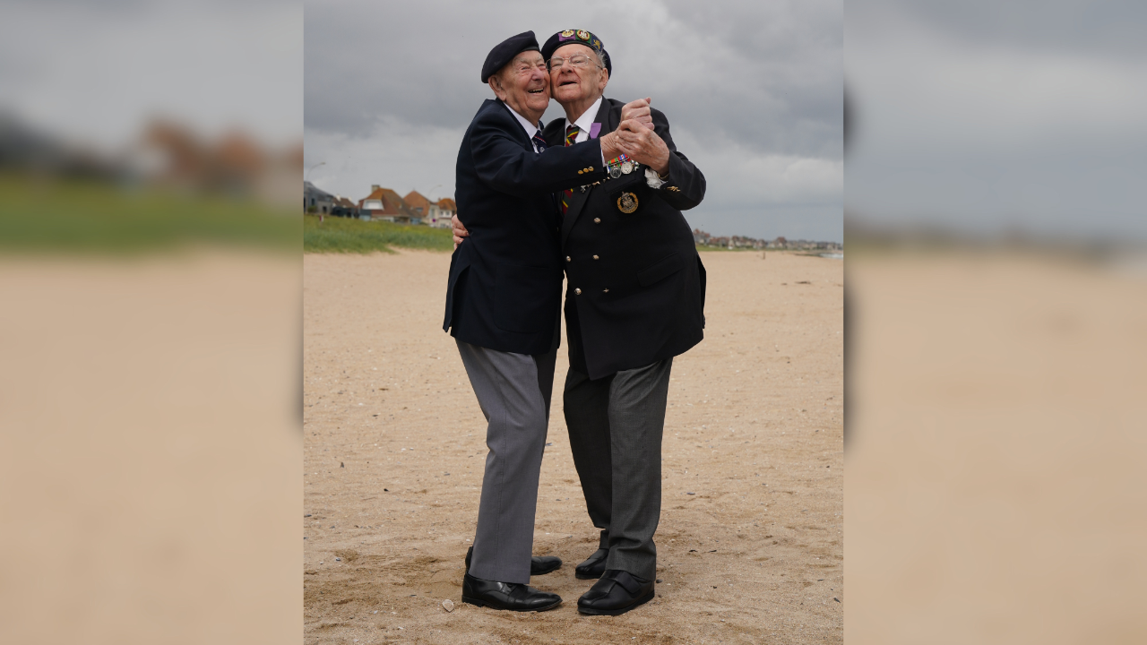 Veterans Ken Hay (right) and Henry Rice (left) on Sword Beach, France, ahead of the 82nd anniversary of D-Day. The lean into each other and hold hands with their arms stretched out. They stand on a sandy beach beneath a grey sky. They are dressed in formal military uniform with medals pinned to their blazers.
