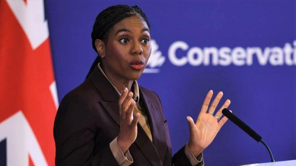 Kemi Badenoch stands at a microphone in front of a Union Jack flag. She is wearing a brown suit.