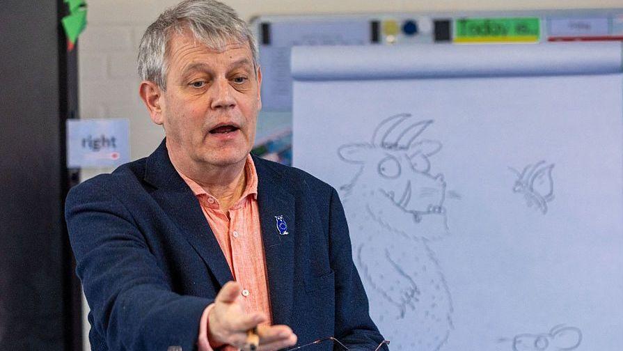 Axel Scheffler, a man with grey hair, wearing a navy jacket over an orange shirt, gestures while speaking in front of a whiteboard displaying a sketch of the Gruffalo.