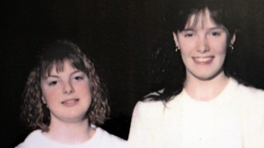 Vintage photograph of two young people standing side by side in light-coloured tops against a dark background. One has shoulder-length curly hair and the other has longer dark hair.