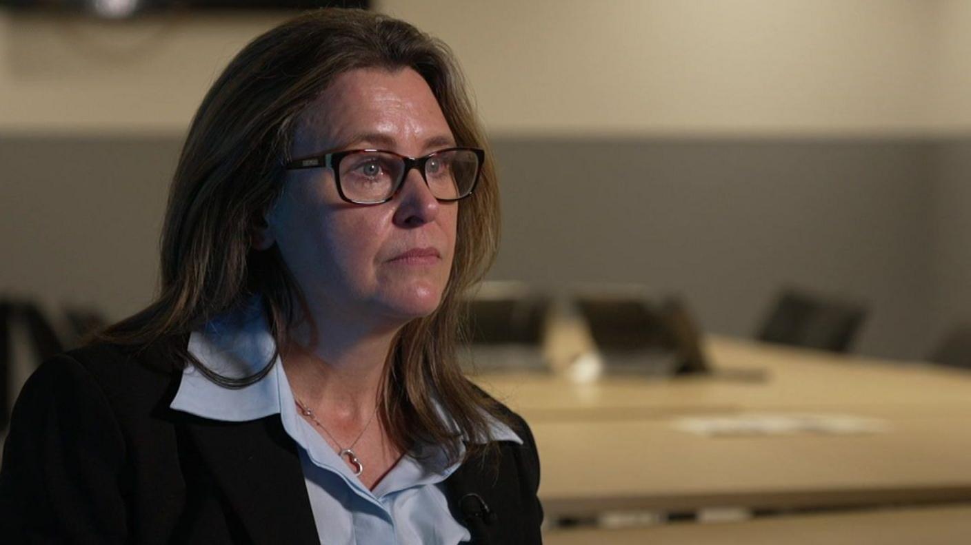Detective Chief Superintendent Emma Bond, wearing a suit, blue shirt and glasses, sitting in an office in front of a large table.