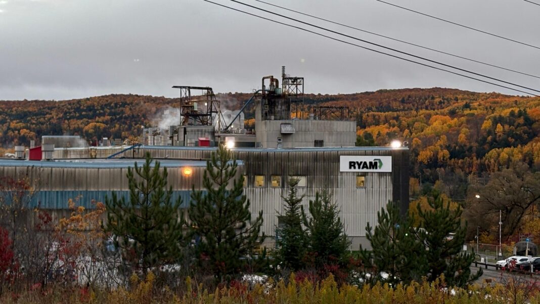 An image showing a part of the RYAM plant in Temsicaming, surrounded by red and orange fall trees. The building is grey with a sign in right corner that features the RYAM logo. Smoke is billowing out the ceiling.