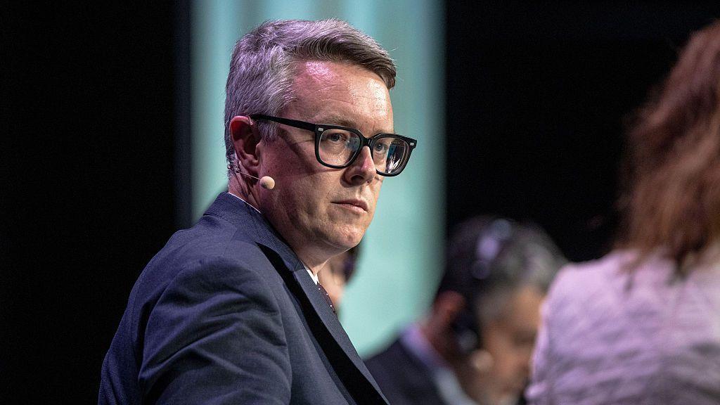 Energy Minister Martin McCluskey, a white man with glasses pictured on stage at an event, with an earpiece and microphone wearing a navy suit.
