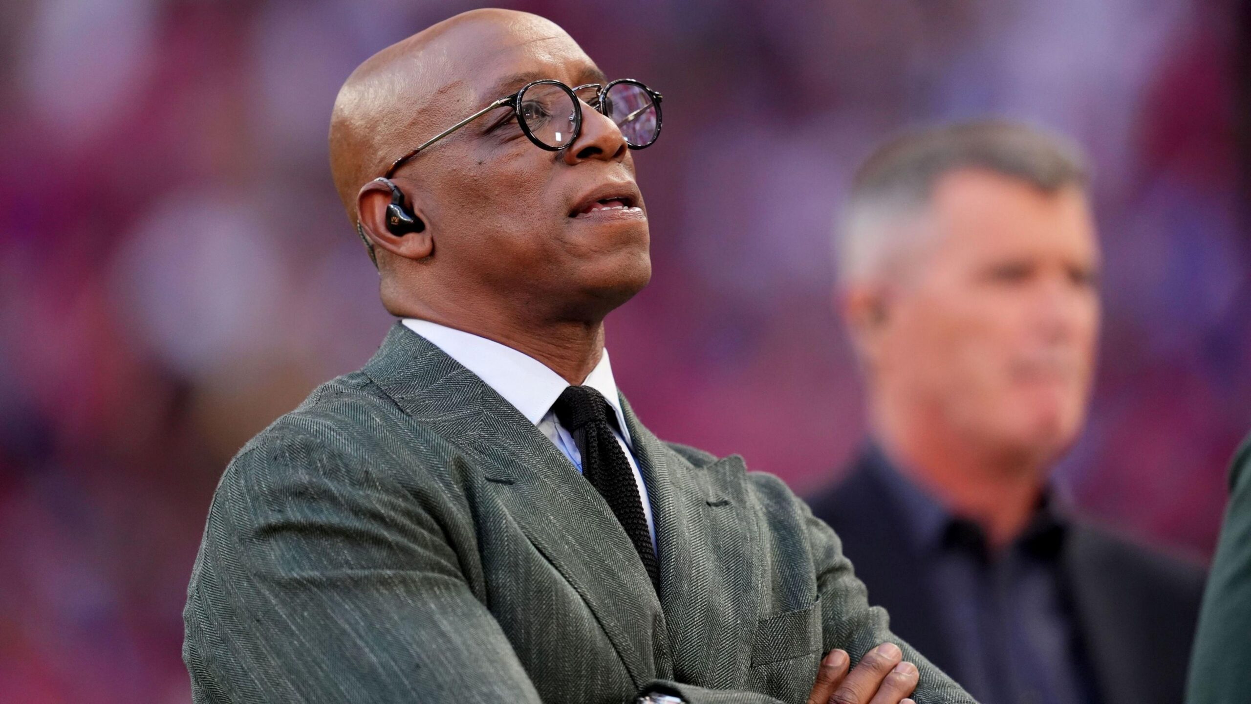 Image of former footballer Ian Wright wearing a grey suit, white shirt and black tie. He is folding his arms and looks like he is watching a football match.