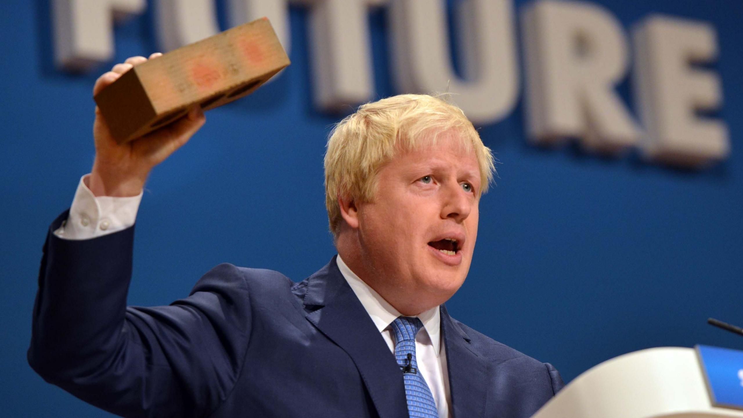 Boris Johnson holds aloft a brick during a speech on the stage at the Tory party conference in 2014. He is wearing a dark blue suit, white shirt and blue patterned tie.