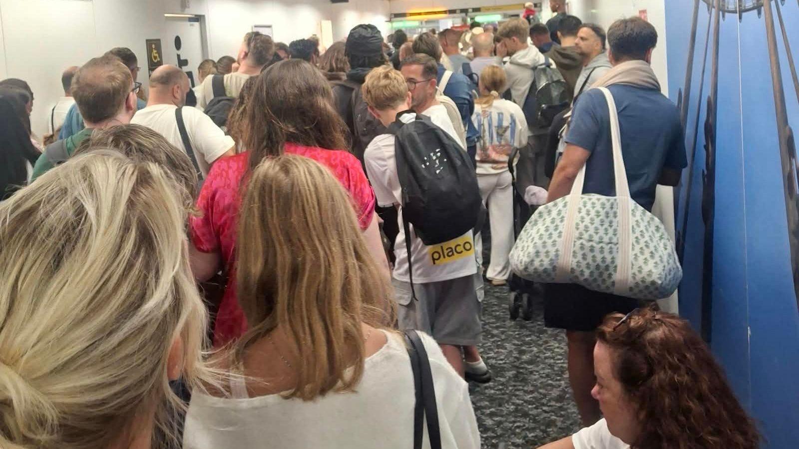 A crowd of people seen from behind queuing in an airport corridor. Most have bags slung over their shoulders.