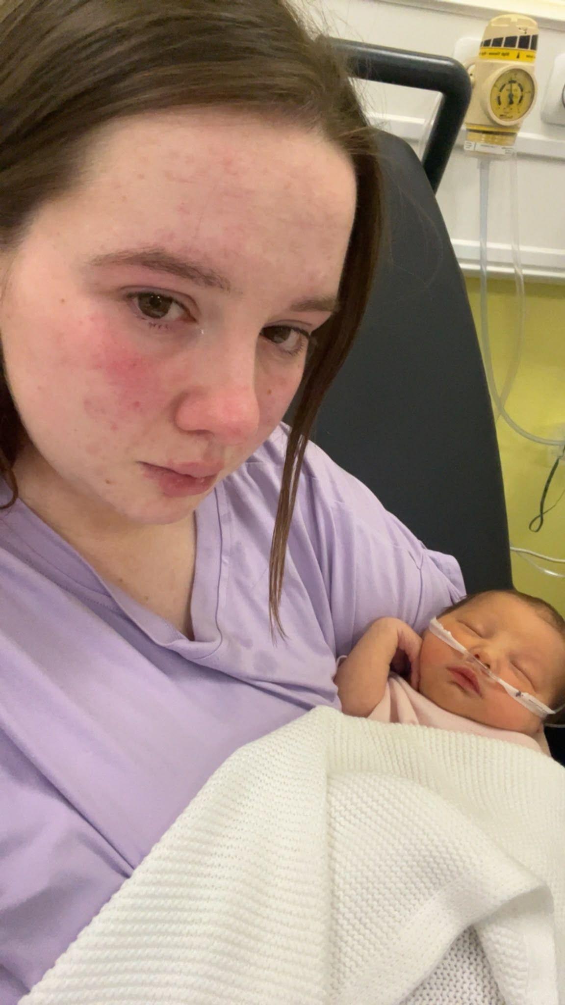 A selfie of Sophie and her daughter Florence. Sophie has brown hair and is holding Florence in her arms. Florence is a newborn baby and is covered by a white blanket. Sophie is looking at the camera and not smiling. Florence has her eyes closed.