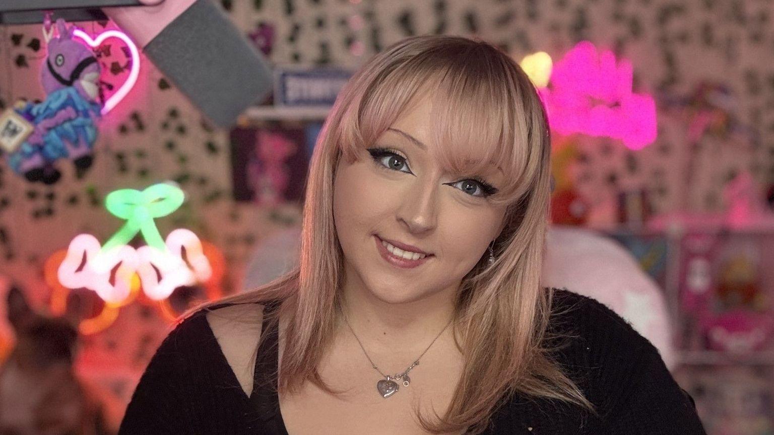 Lisha sits in a pink gaming chair and faces the camera smiling. In the background are flourescent signs and a patterned backdrop. She has light link hair and wears a black jumper and silver necklace.
