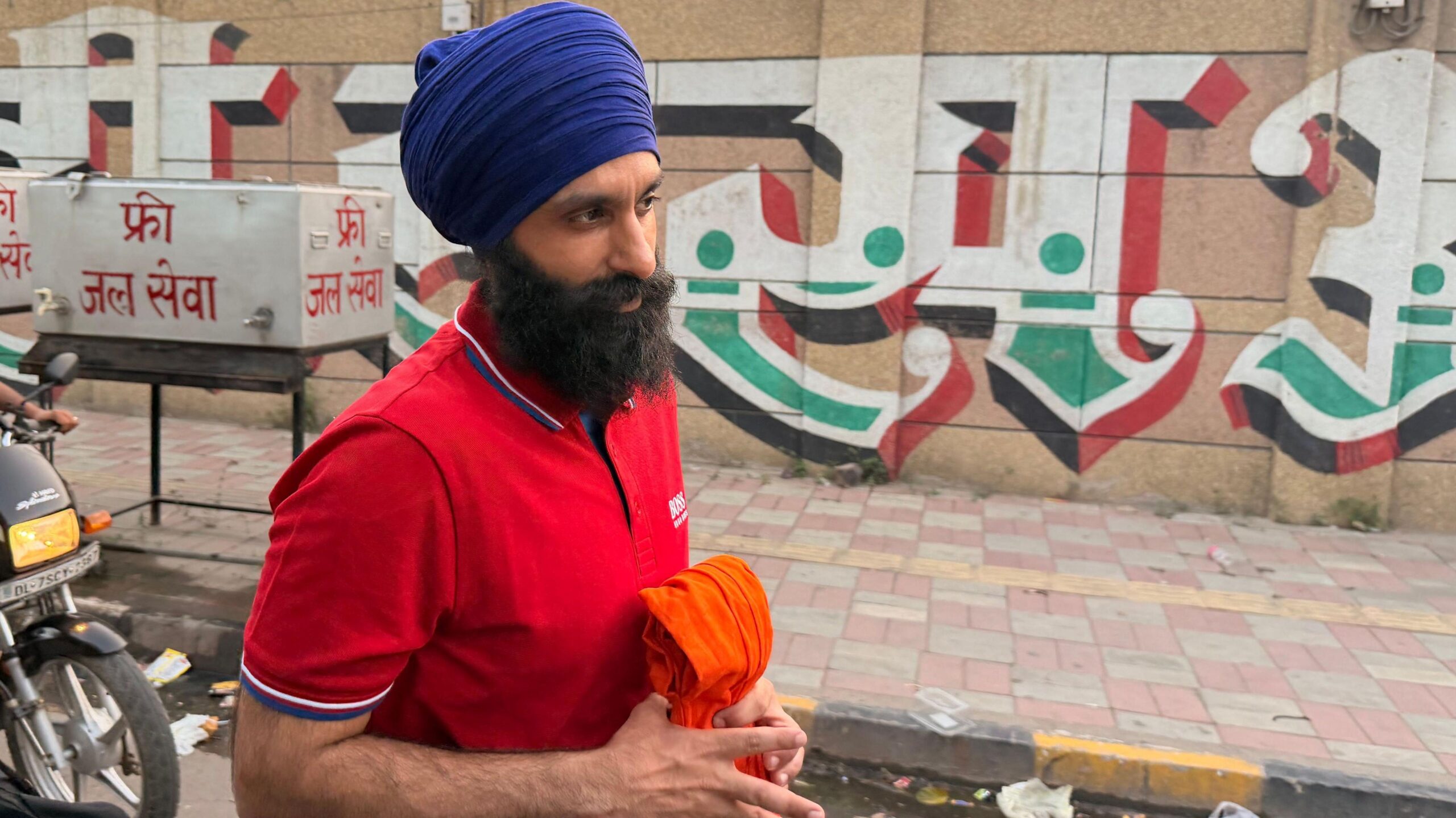 Jagtar Singh Johal looking off camera. He is wearing a red polo shirt with the word 'Boss' on the upper right chest. He is wearing a purple turban around his head and has a long, dark beard. He is clutching an orange garment in his hand and is walking on a street with a motorbike in the background.