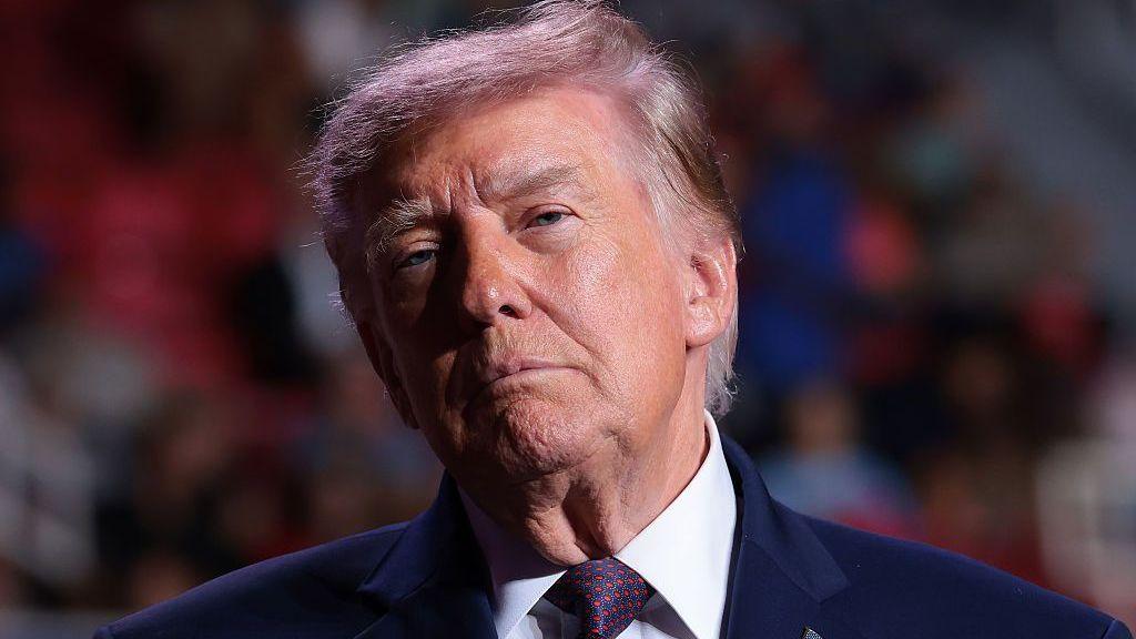 Donald Trump stands in front of a blurred crowd.
