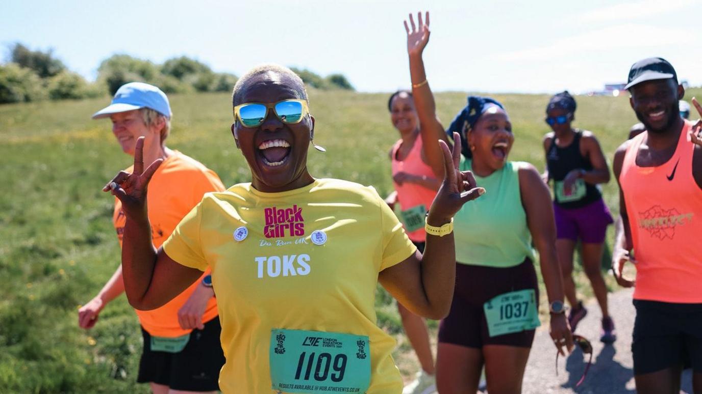 A female runner grins at the camera as she holds both hands up in a peace sign. There are several other runners behind her who are also smiling.