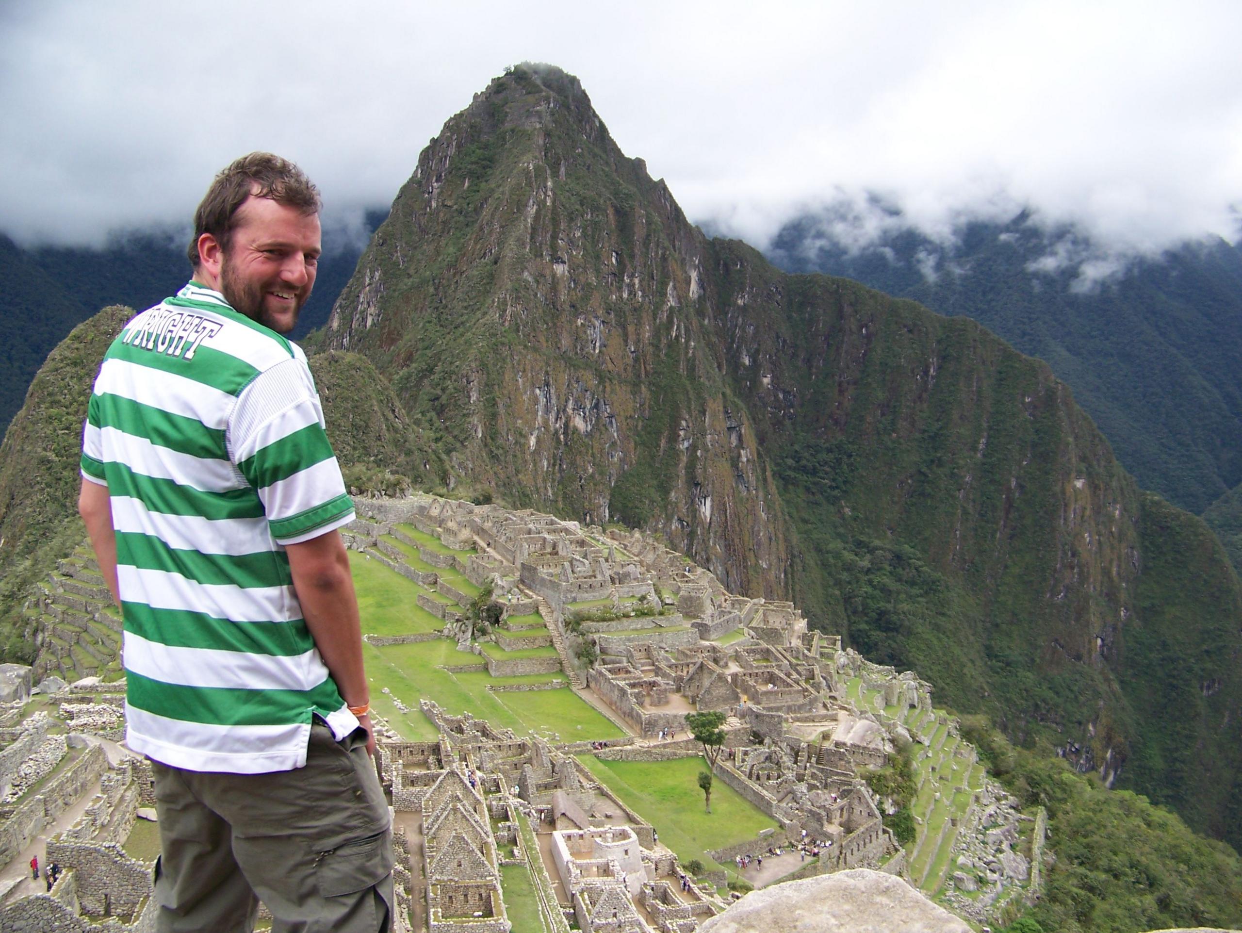 A man in the strip turns slightly to the camera with his back to the front, the citadel of Machu Picchu laid out in front of him.