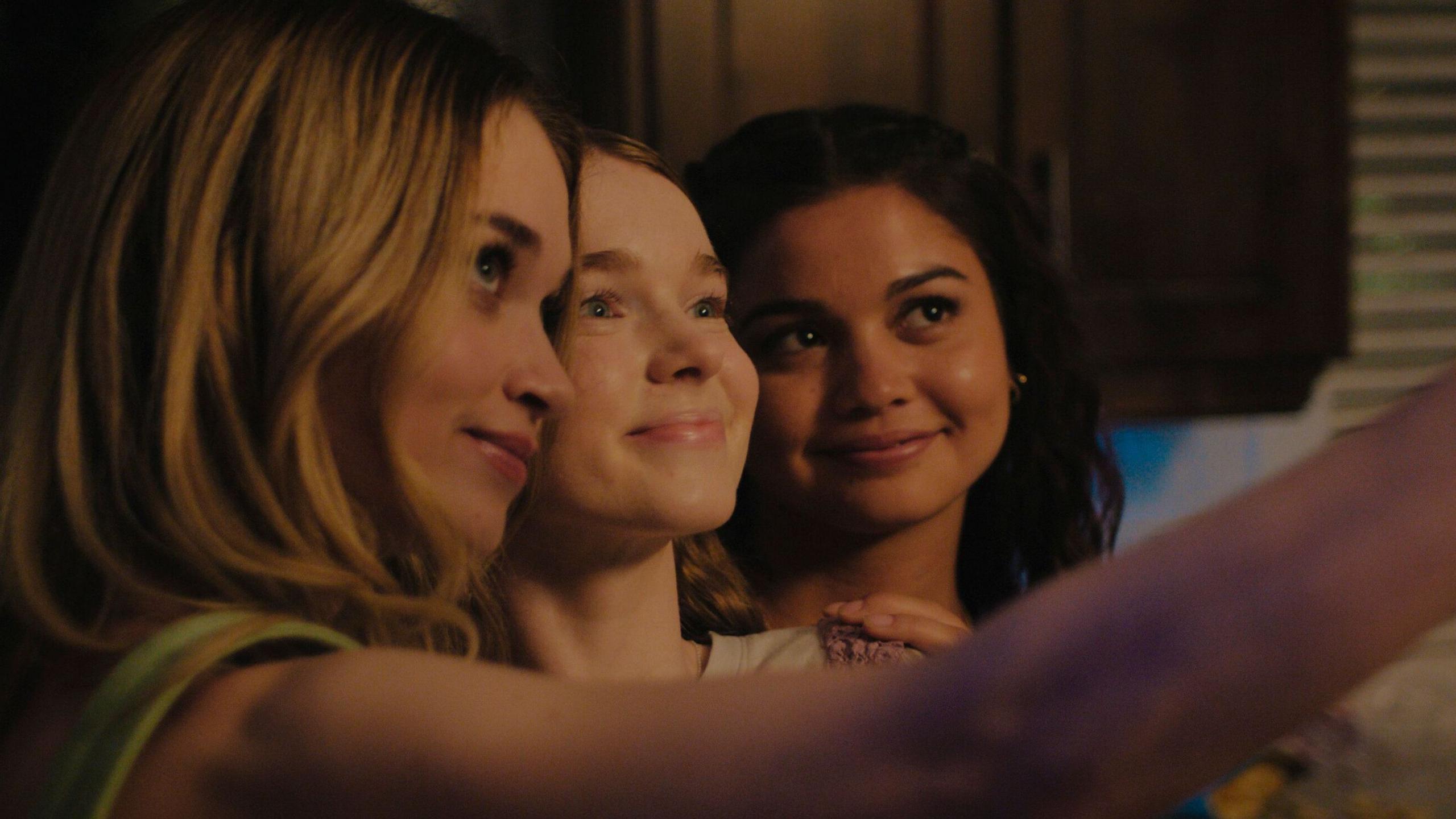 Still from Carrie shows her standing between Chris Hargensen and Sue Snell in a dimly lit kitchen. The three girls are huddled close together, smiling, and Chris's arm is outstretched, framing a selfie of the group.