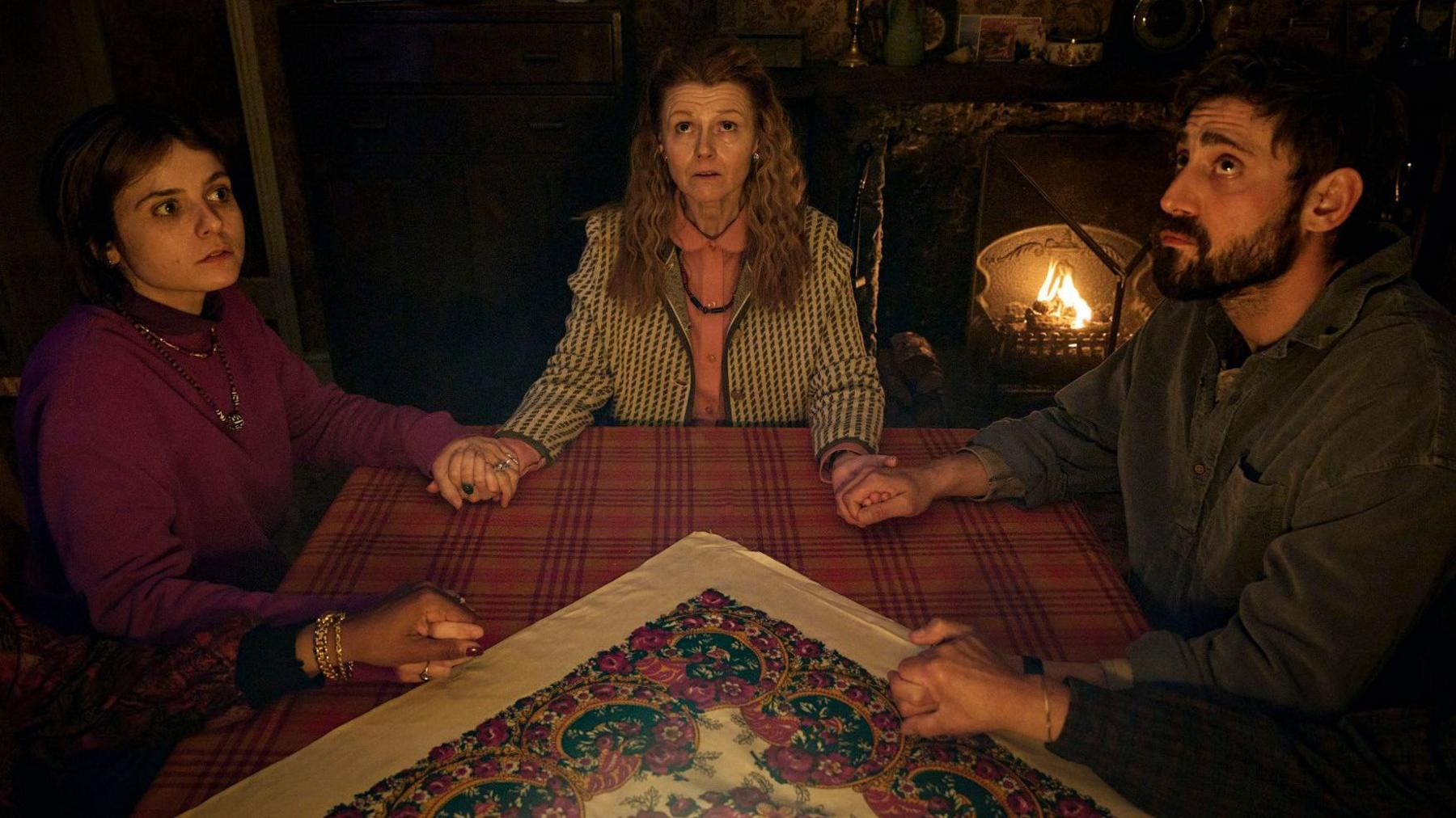 A woman with long dark brown hair and a burgundy top sits at a table in a house and is holding hands with a woman with long curly red hair, a peach-coloured blouse and a dogtooth coat. A man with dark brown hair and a beard is also seated at the table and is also linking hands. The hand of another person who is out of shot can also just be seen on the table. The three people look concerned as their eyes are focused above or seemingly in the distance. A patterned tablecloth covers the table.