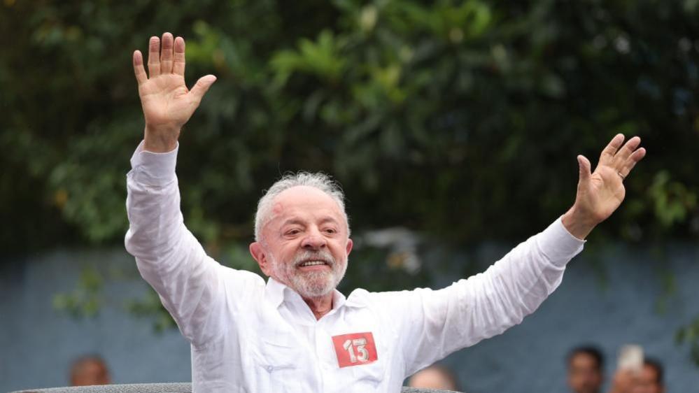 Luiz Inacio Lula da Silva greets supporters after casting his ballot. He is smiling and waving as he stands through the sunroof of a car.