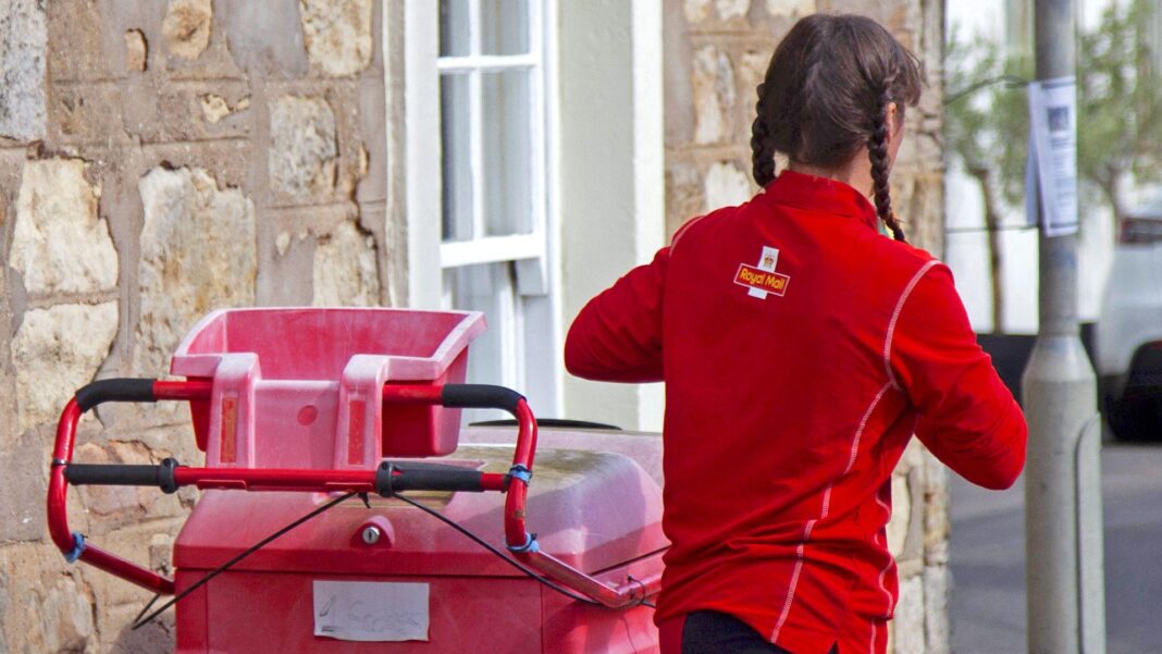 A postwoman seen from behind, wearing a Royal Mail top and standing beside a mail trolley on a residential road