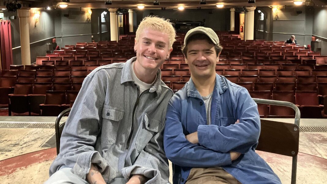 Gaten Matarazzo on life after Stranger Things as he makes West End debut Travis and Gaten sit next to eachother on stage. Gaten (right) wears a blue jacket, striped top, khaki trousers and a cap. Travis has blonde hair, wears a blue cord shirt with a white t-shirt underneath.