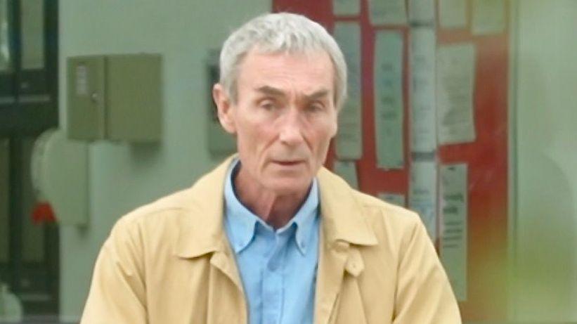 A man outside a court building on his phone. He wears a yellow coat and blue shirt. He has short grey hair.