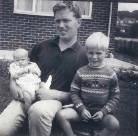 A black-and-white family photograph taken outdoors in front of a brick house. Rex is seated on a chair holding Rachel as a baby on a lap. Ian is sitting beside Rex, wearing a patterned knitted jumper and shorts. A window, garden area and fencing are visible in the background. The three are positioned close together in the foreground.