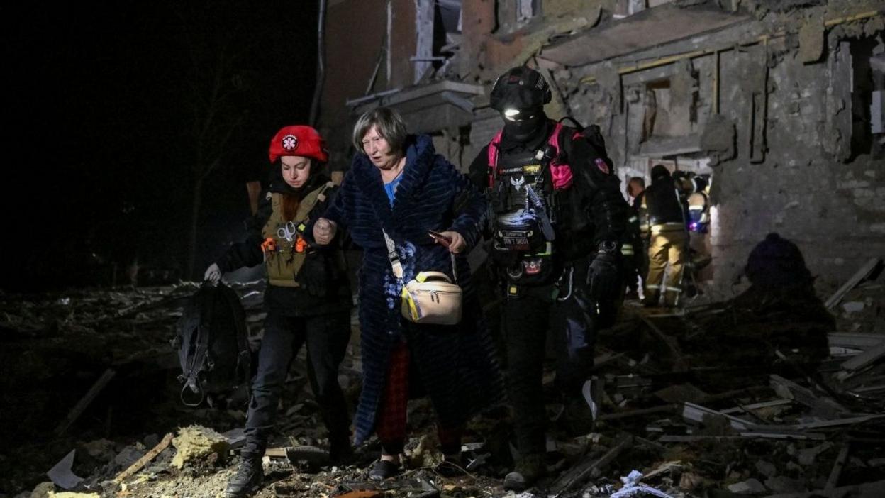 Wide shot showing a middle-aged woman walking  away from a residential building that has been damaged by Russian strikes at night. She is holding the hands of two rescue workers who are helping her walk over the rubble.