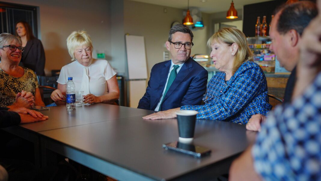 Andy Burnham, in a blue suit and glasses, sat with people including a woman with blonde hair, in a blue checked jacket