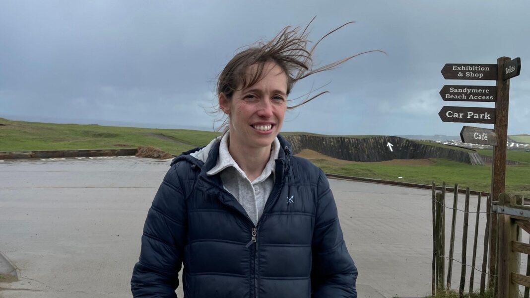 Devon and Cornwall battening down the hatches ahead of Storm Austen A woman wearing a coat stands with exposed grassy cliffs behind her. Her hair is blowing in the wind.