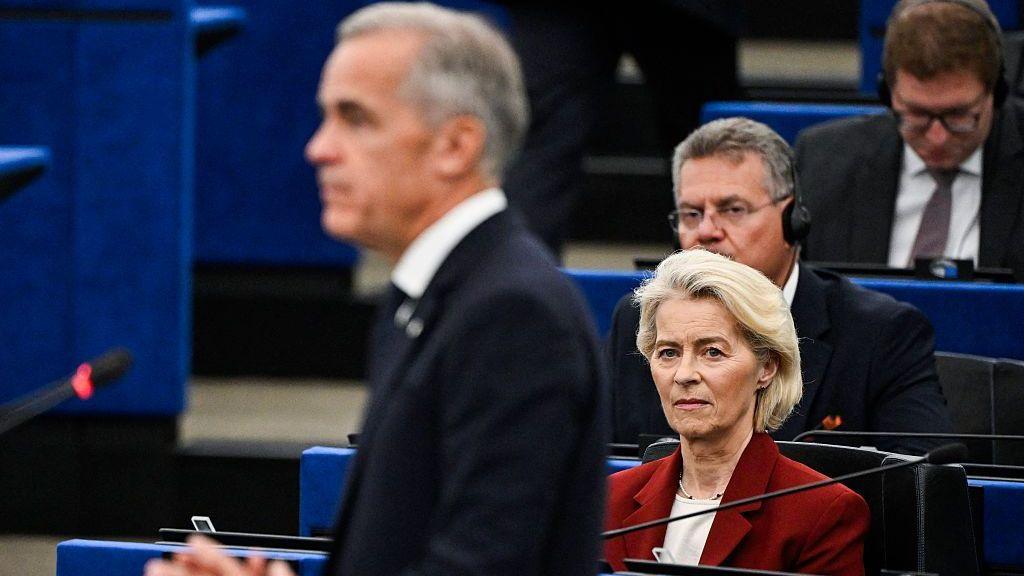 An image showing European Commission President Ursula von der Leyen looking at an out of focus Mark Carney as he delivers a speech to the European Parliament. She has short blonde hair and is wearing a maroon jacket.