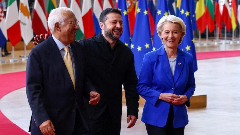 António Costa, Volodymyr Zelensky and Ursula Von der Leyen laughing as they walk, with many country flags hanging behind them.