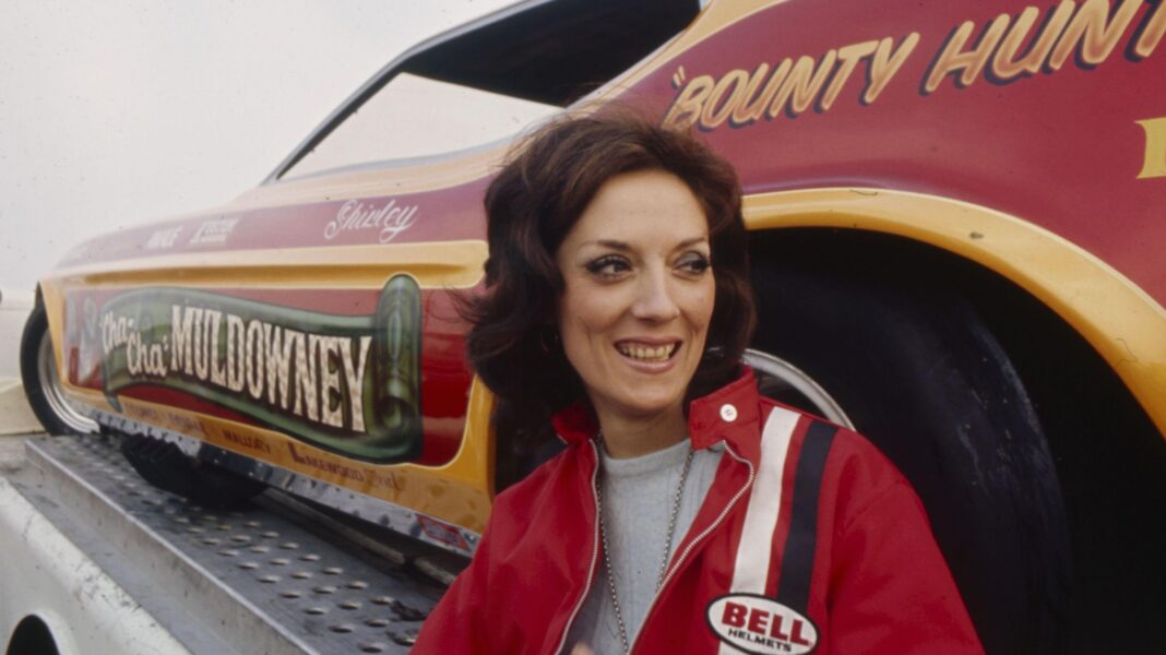 Drag racing trailblazer Shirley Muldowney pictured at the National Hot Rod Association (NHRA) Winternationals at Pomona Raceway in 1970. She is wearing a red jacket and smiling, in front of her car, which is red with 