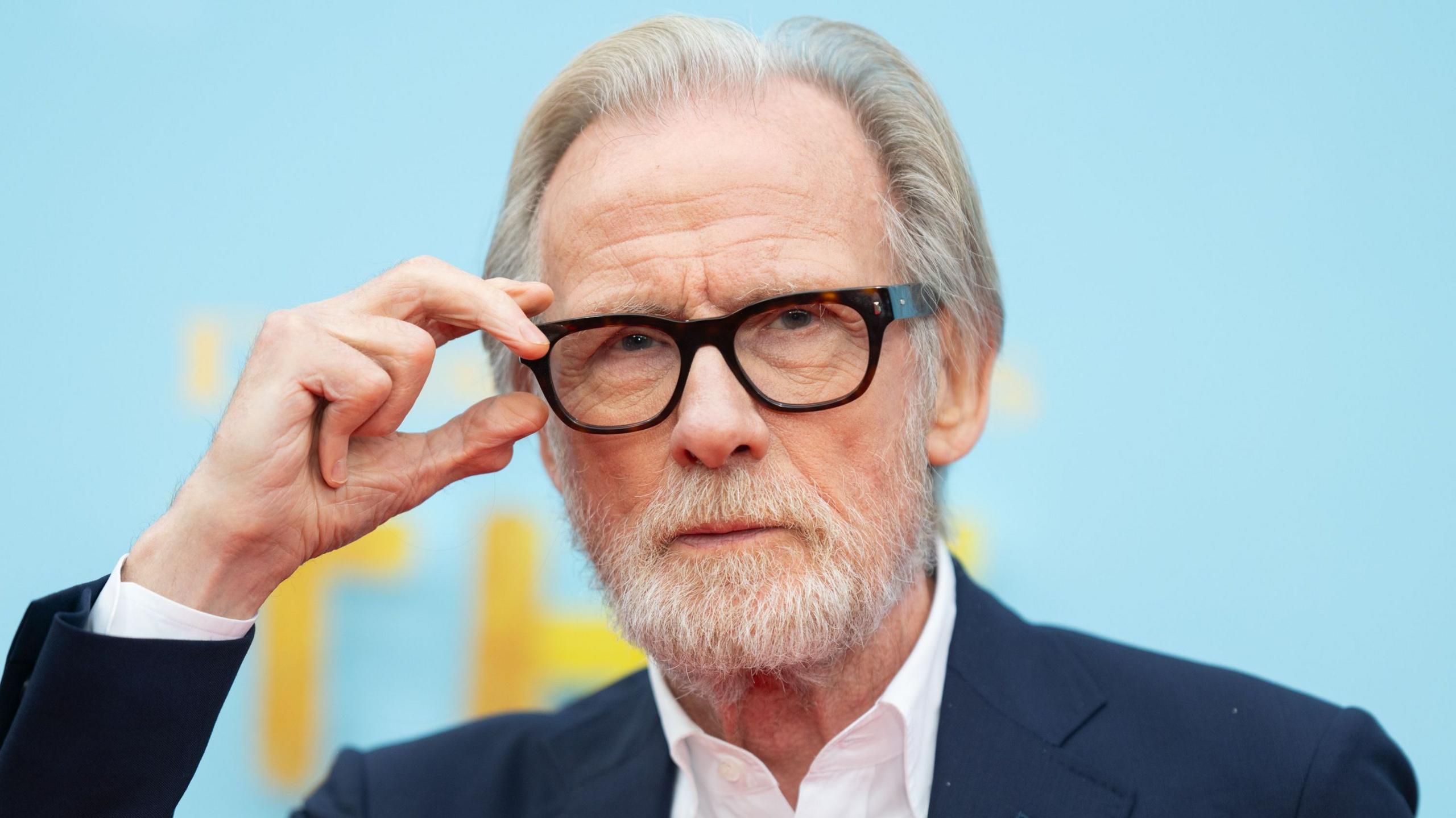 Bill Nighy in a dark suit with a white open necked shirt, with one hand on his black glasses