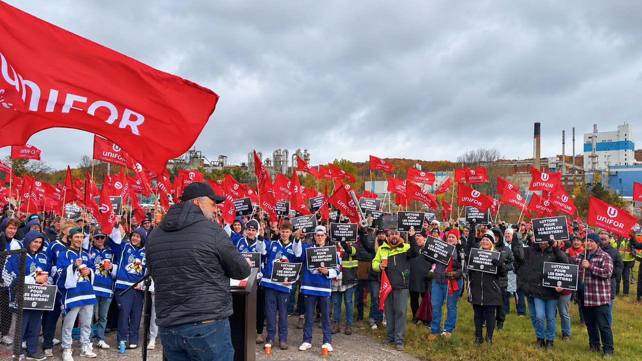 Témiscaming residents hold a union-led rally to support the mill's workers - participants hold red union flags and some have uniform blue jackets - a man wearing a cap addresses them from a podium.
