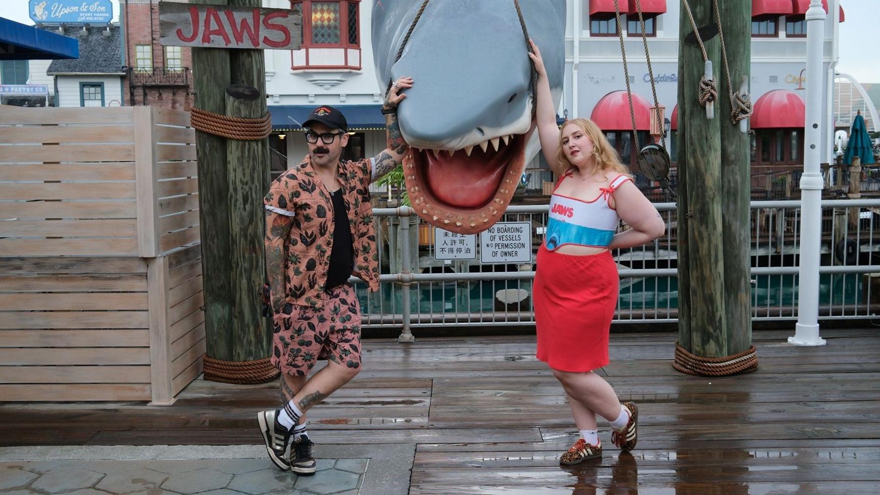 Gav and Honey posing in front of a shark near a Jaws sign. Gav is in a tropical two piece set, while Honey is wearing a Jaws top and a red skirt.