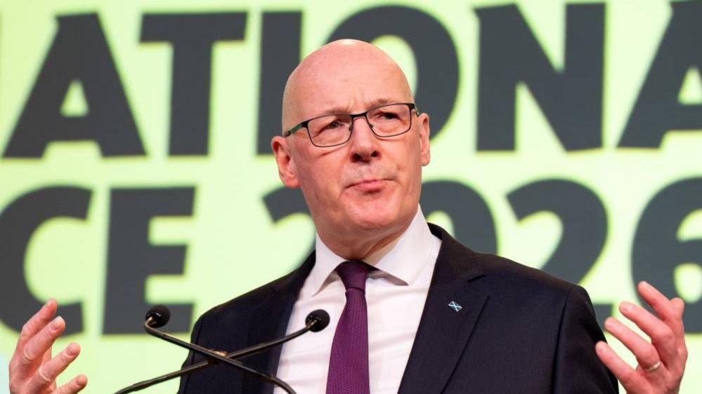 John Swinney, a bald man with glasses wearing a black suit and purple tie with a saltire pin on it. He has his hands outstretched beside him and two microphones in front of him. He is mid speech. Behind him is a yellow sign with black writing.