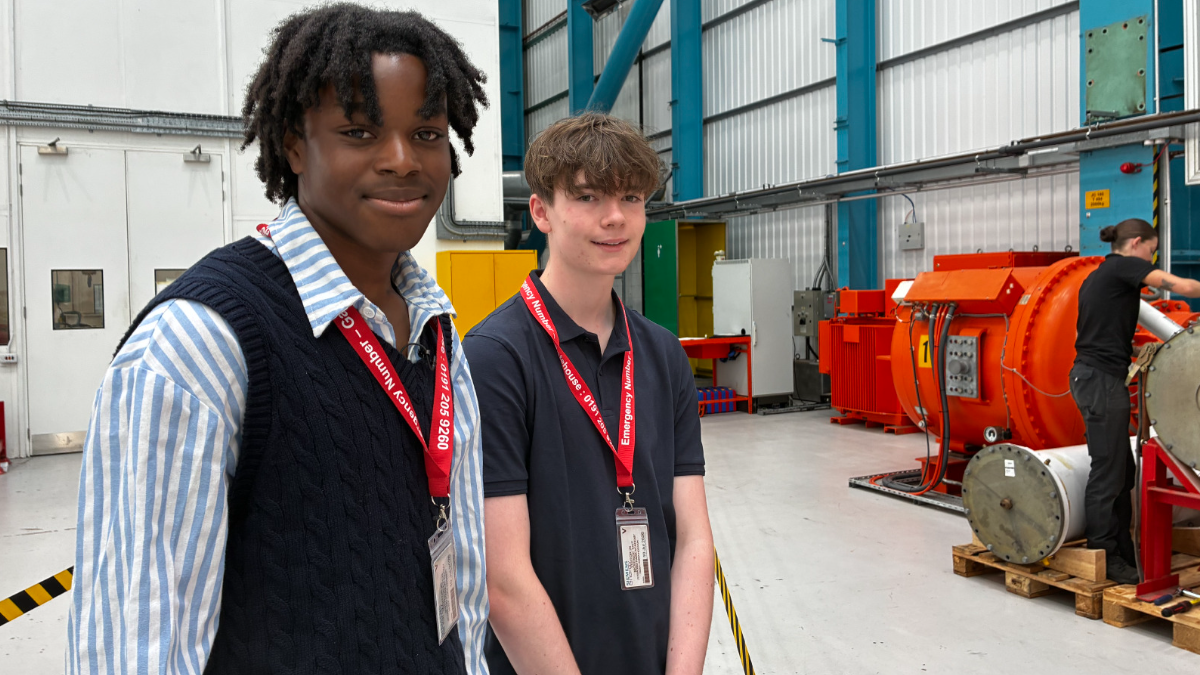 Javaris and McKenzie are pictured wearing visitor lanyards inside an industrial workshop beside large orange machinery. The spacious facility has high ceilings, blue steel supports and production equipment in the background, with another person working near the machinery.