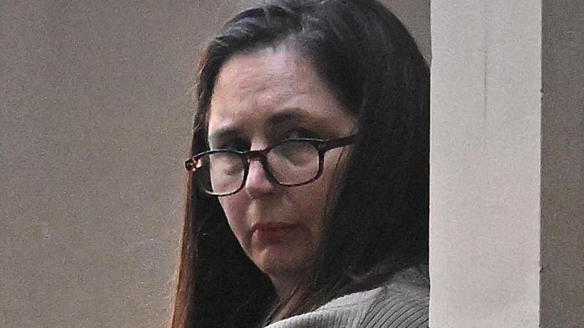 Erin Patterson looks at the camera, in a grey jumper wearing black glasses. Her brown hair is down.