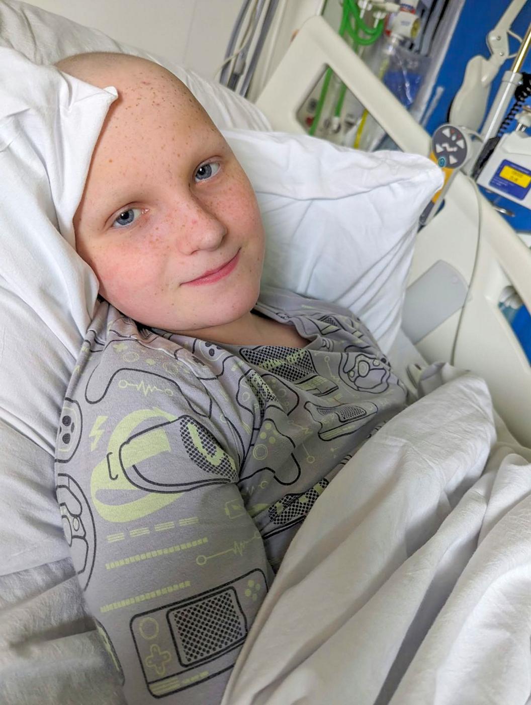 A boy of around 13 years old lies in a hospital bed looking directly at the camera. He has no hair or eyebrows and is wearing a pyjamma top.