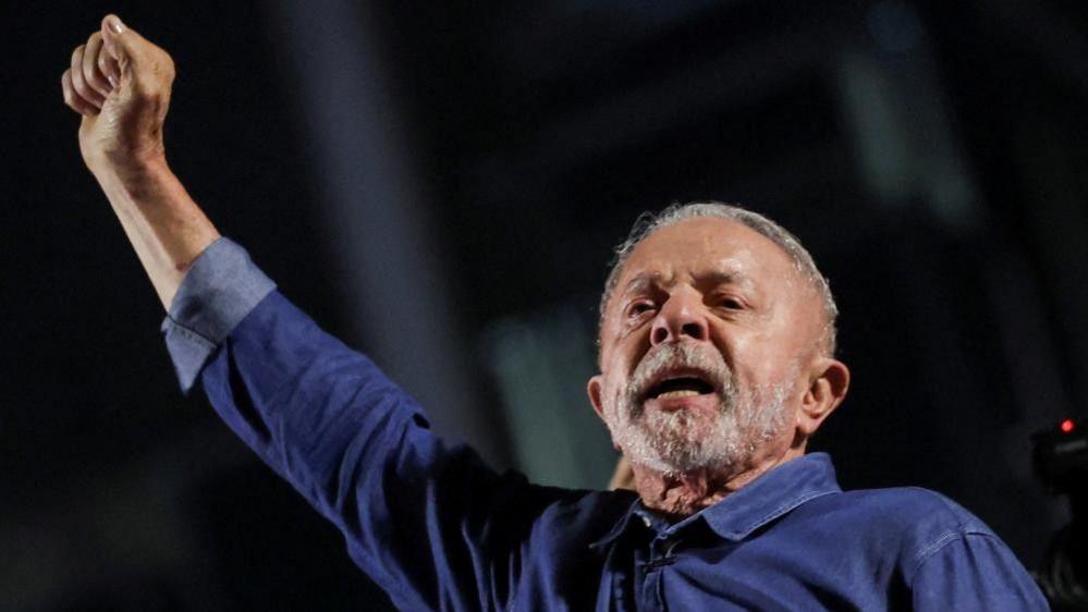 President Lula stretches his fisted right arm as he addresses supporters. He's wearing a blue short and has a grey beard and short hair.