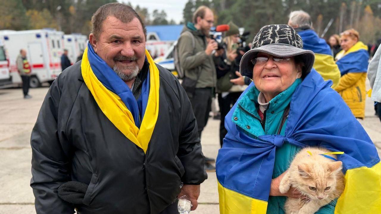 An older man, wearing a black bomber jacket, smiles as he is pictured next to his wife who is wearing a turquoise rai jacket and a bucket hat, and is holding a cat