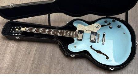 A pale blue electric guitar in an open black case, lying on a grey wooden floor