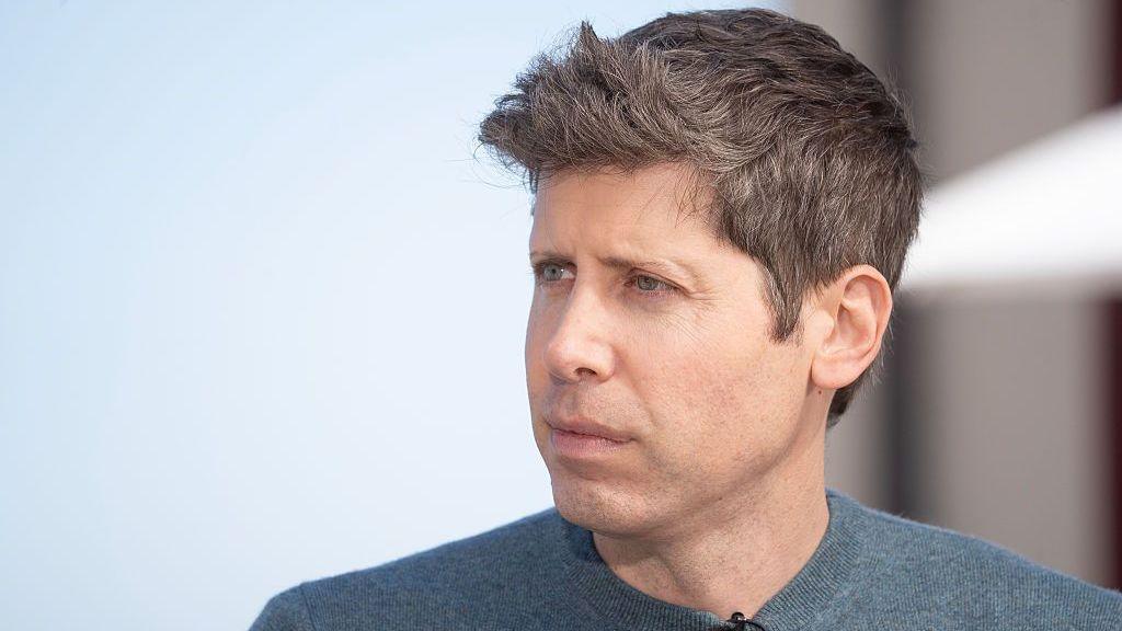 Sam is wearing a grey-blue sweater and looks serious in a close up image.