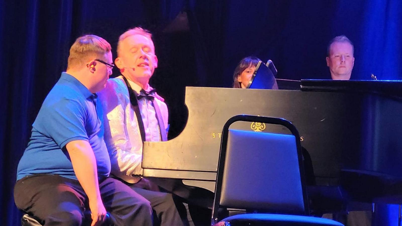 Stanley in a white blazer with a black bow tie playing a piano while sat next to one of his students.