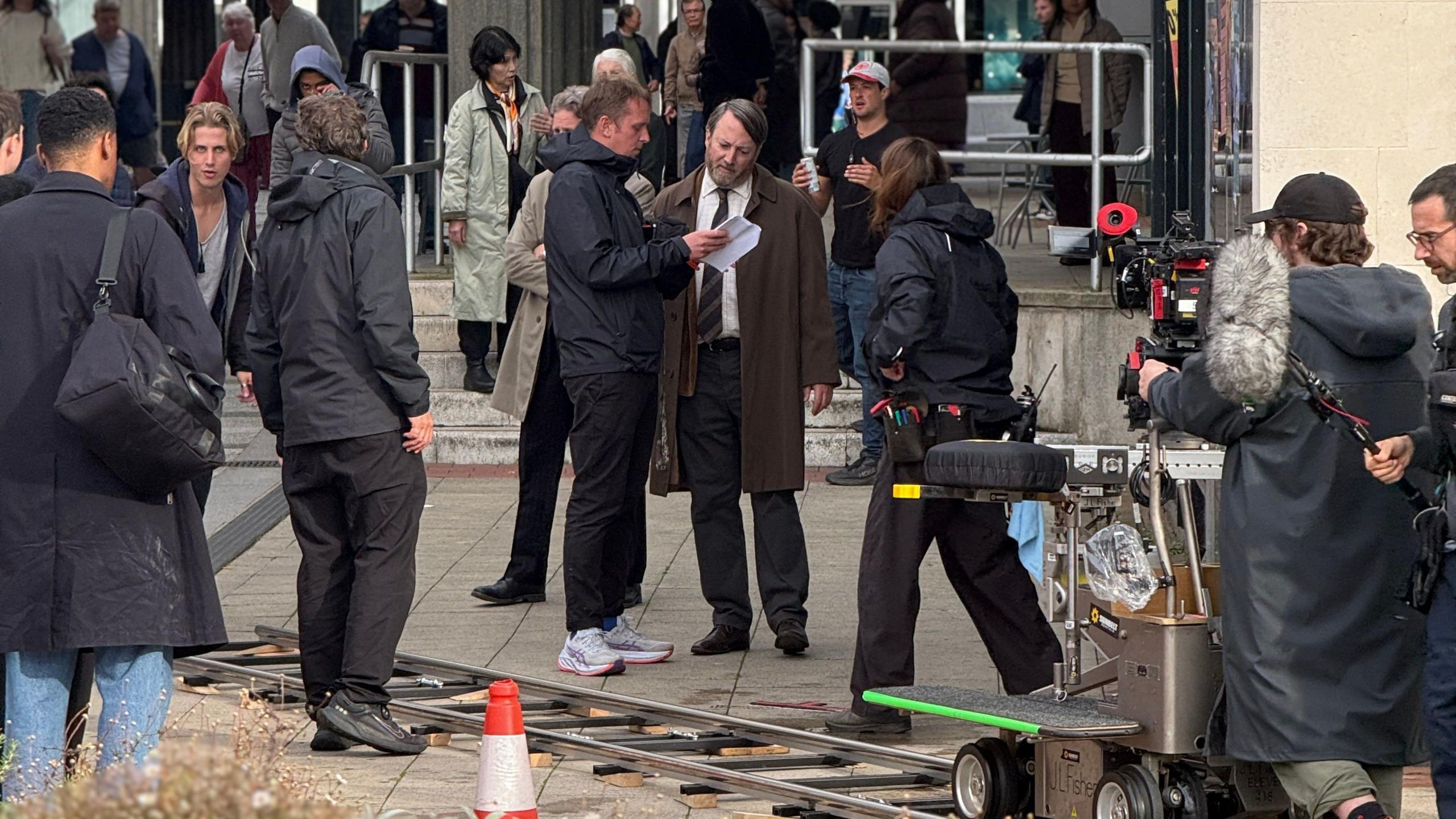 A busy street full of extras and film crews, David Mitchell is stood in the middle reading a script with a man dressed in black