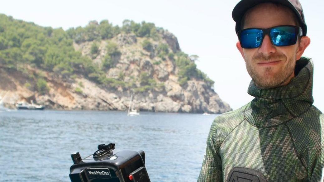 A man in a wetsuit, baseball cap and sunglasses. he is holding a camera. Behind him you can see the sea, small boats and rocky coastline.