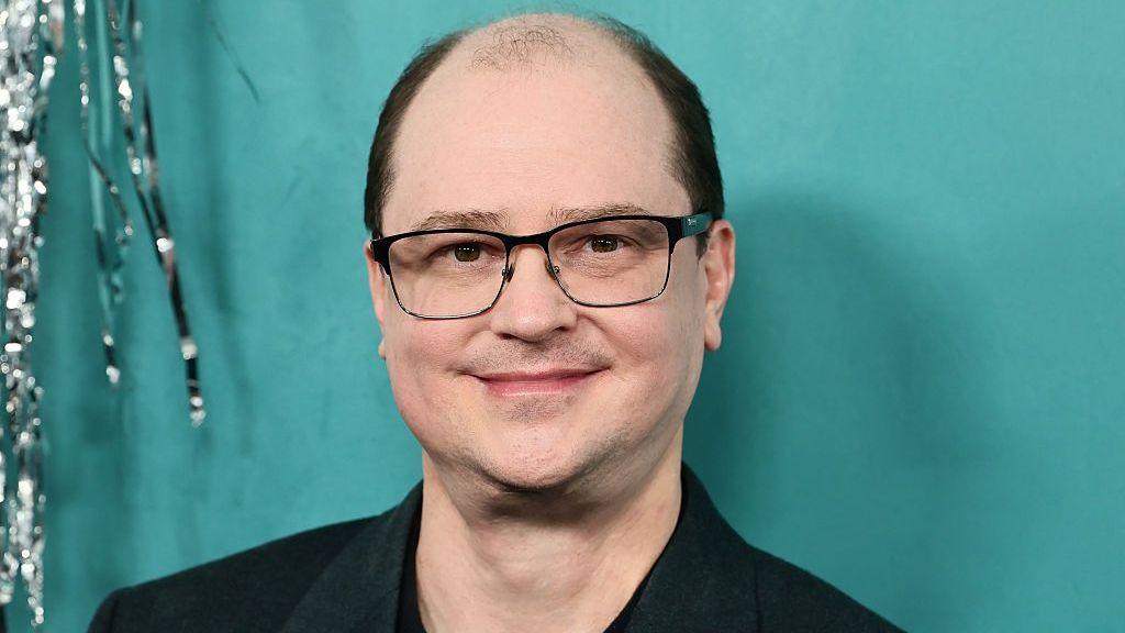 A man wearing glasses and a black suit jacket stands in front of a teal coloured background, posing for photographers. A tinsel-style streamer can be seen dangling down behind him.
