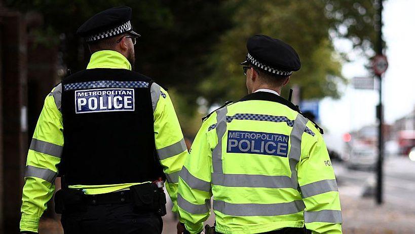 Two Met Police officers face away from the camera as they walk down a street in London. Both are wearing hats and hi-viz jackets. One on the left wheres a black vest over his jacket.