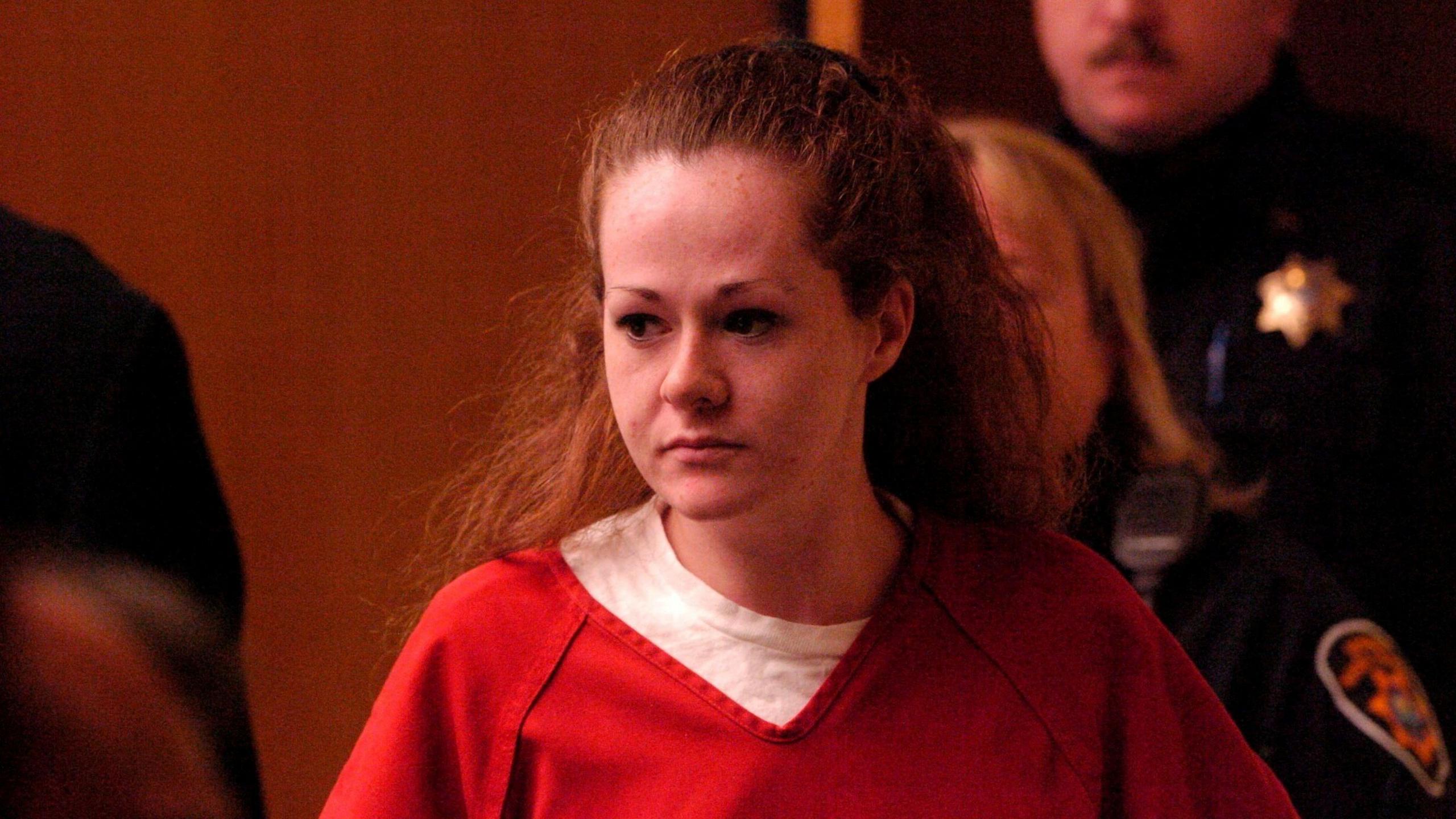 Christa Pike is pictured looking sombre in red prison scrubs over a white t-shirt, her long auburn hair tied up halfway and uniformed guards visible behind her