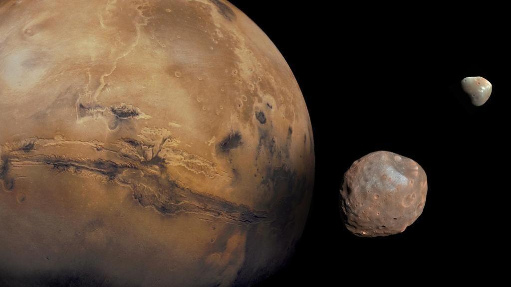 Mars fills most of the scene - it is orange and its surface is pitted with craters and canyons. Next to Mars, and against the blackness of space, there are its two small, irregularly shaped moons. In the foreground, the larger moon Phobos is pale red and covered in craters. In the distance, the smaller moon, Deimos, is grey and orange with an irregular shape.