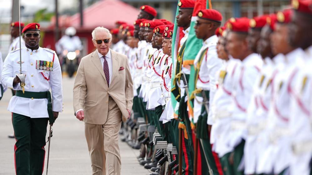 King Charles III, in a light brown suit, observes a guard of honour. He walks along a line of members of the Guyana Defence Force, who wear white shorts with military honours on them, and green trousers.