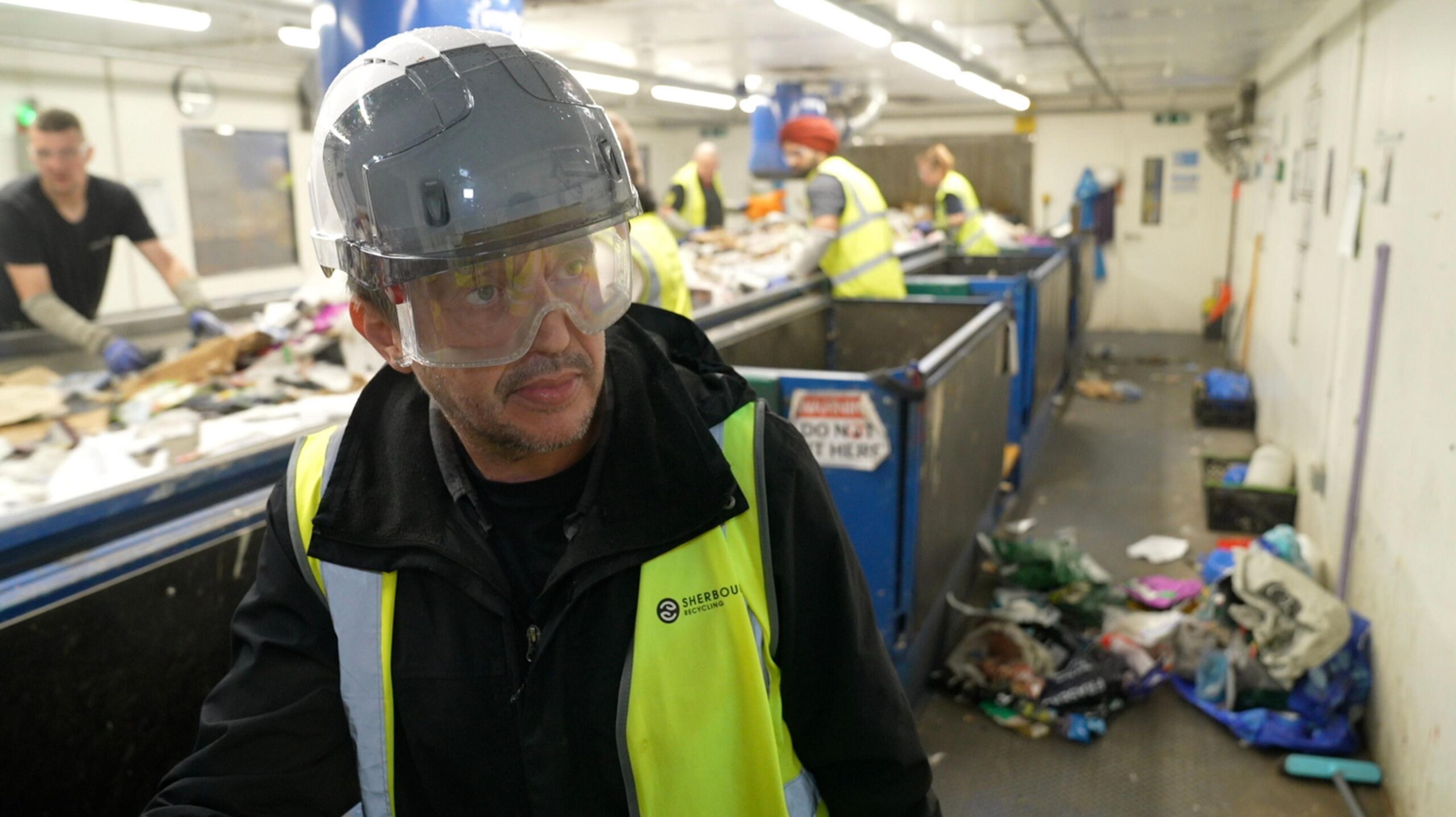A worker wearing a high-visibility vest, safety helmet and protective goggles stands beside a waste-sorting conveyor belt inside a recycling facility. In the background, several other workers in high-visibility clothing sort mixed waste by hand along the conveyor line. Overhead fluorescent lights illuminate the industrial workspace, where piles of recyclable materials and waste are visible on the conveyor and on the floor.