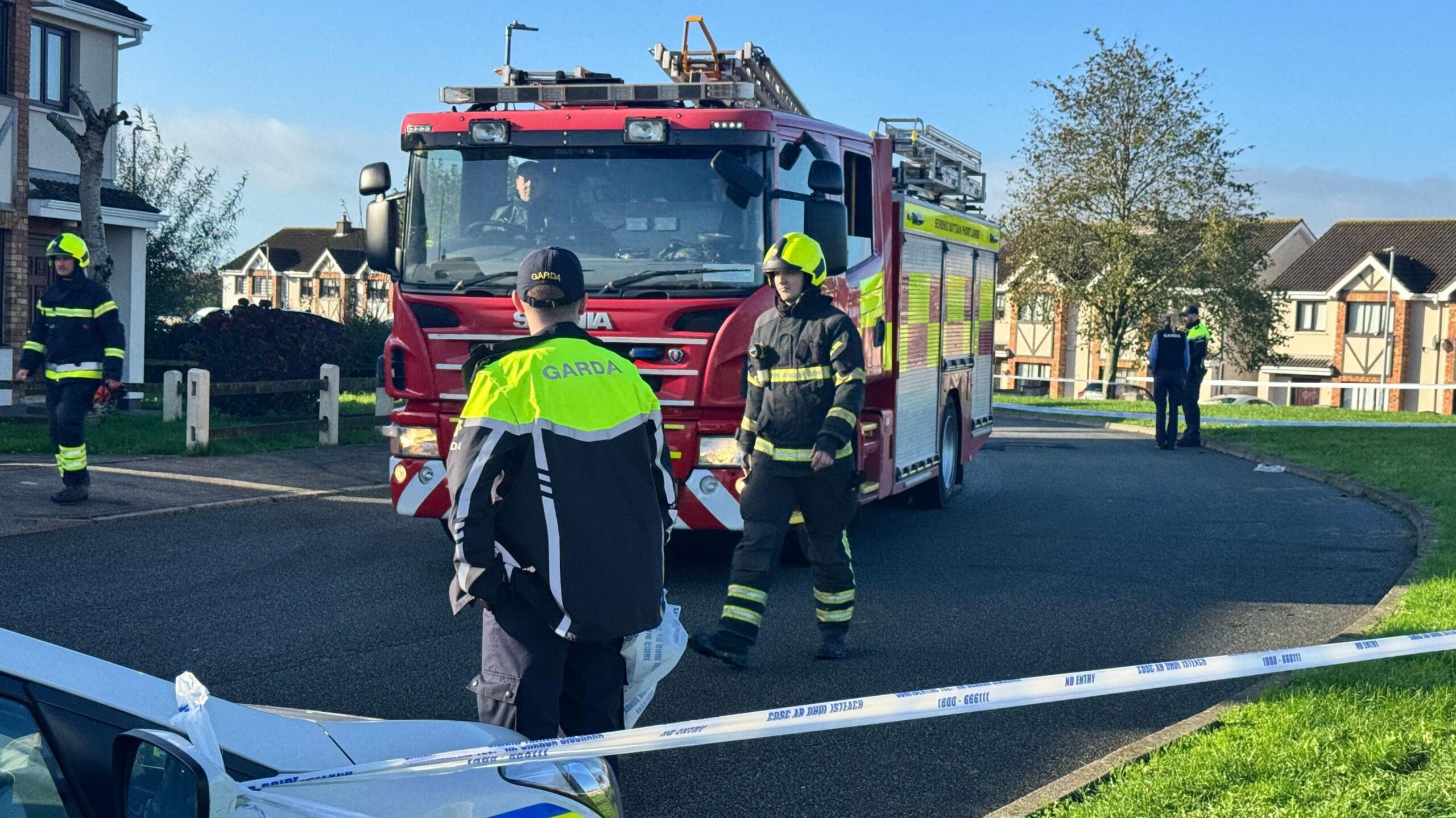 Firefighers and uniformed gardaí (Irish police) at the scene of a fatal house fire in a housing estate in Waterford.  A red fire engine is parked on the road in the middle of the photo.  The street is lined by large semi-detached houses.  A blue and white tape cordons off the road.